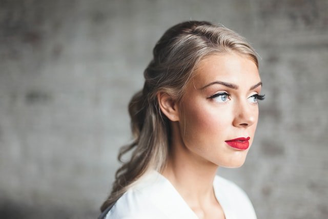 blondynka ze spiętymi włosami i czerwonymi ustami spogląda w dal na tle szarej ściany w białej bluzce