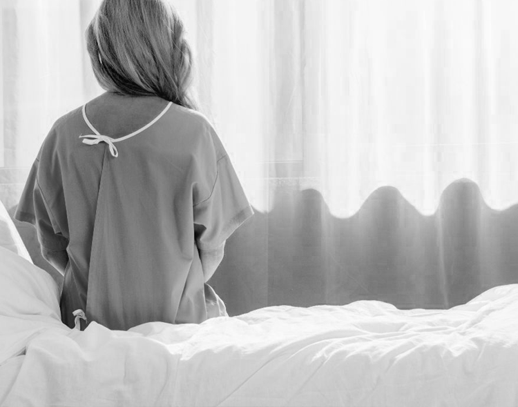 kobieta w szpitalnej koszuli siedzi tyłem na skraju łóżka i patrzy przez okno