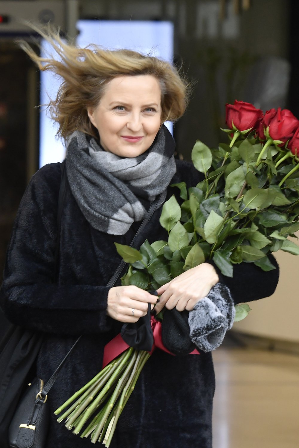 Izabela Kuna w czarnym płaszczu i z naręczem czerwonym róż