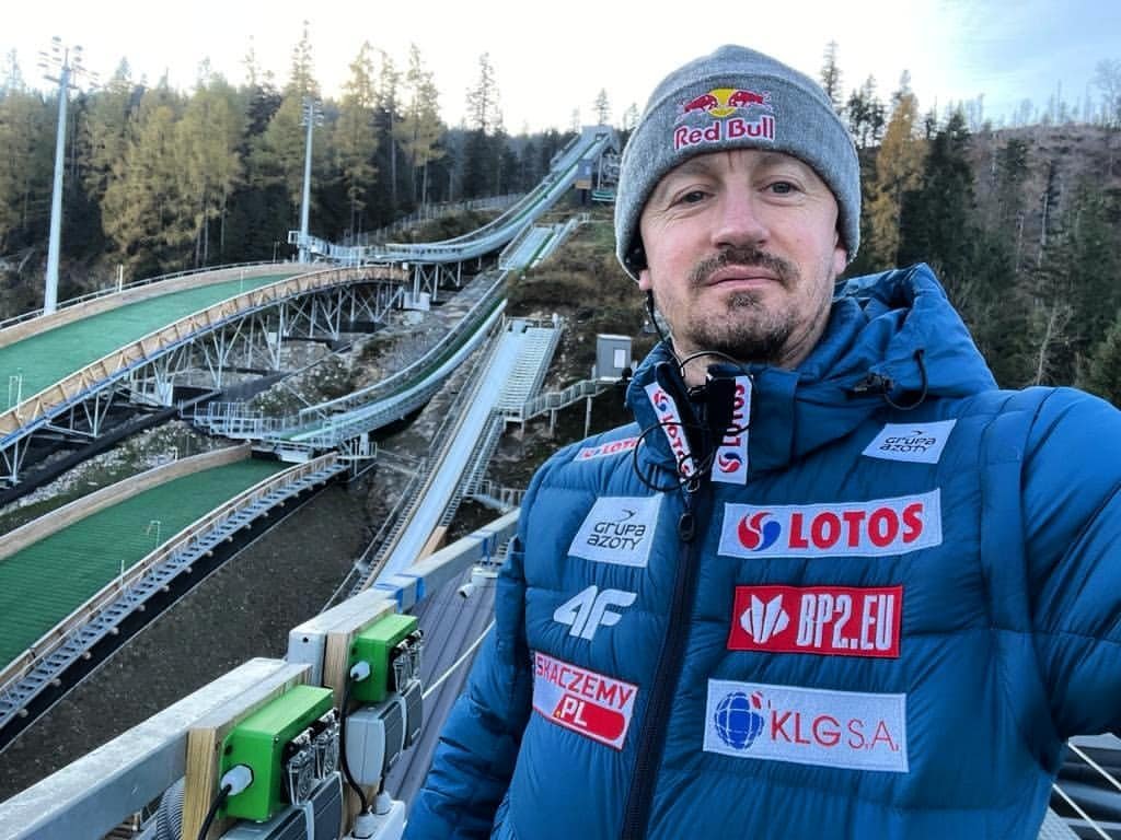 Adam Małysz na skoczni w szarej czapce i niebieskiej kurtce