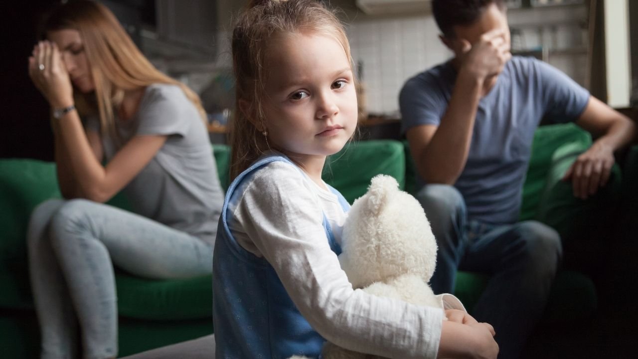 Na pierwszym planie smutna dziewczynka z białym misiem a w tle rodzice siedzący na kanapie