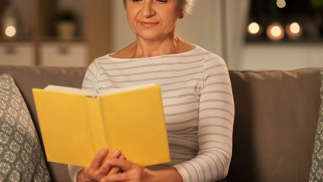 starsza kobieta siedzi na kanapie i czyta żółtą książkę