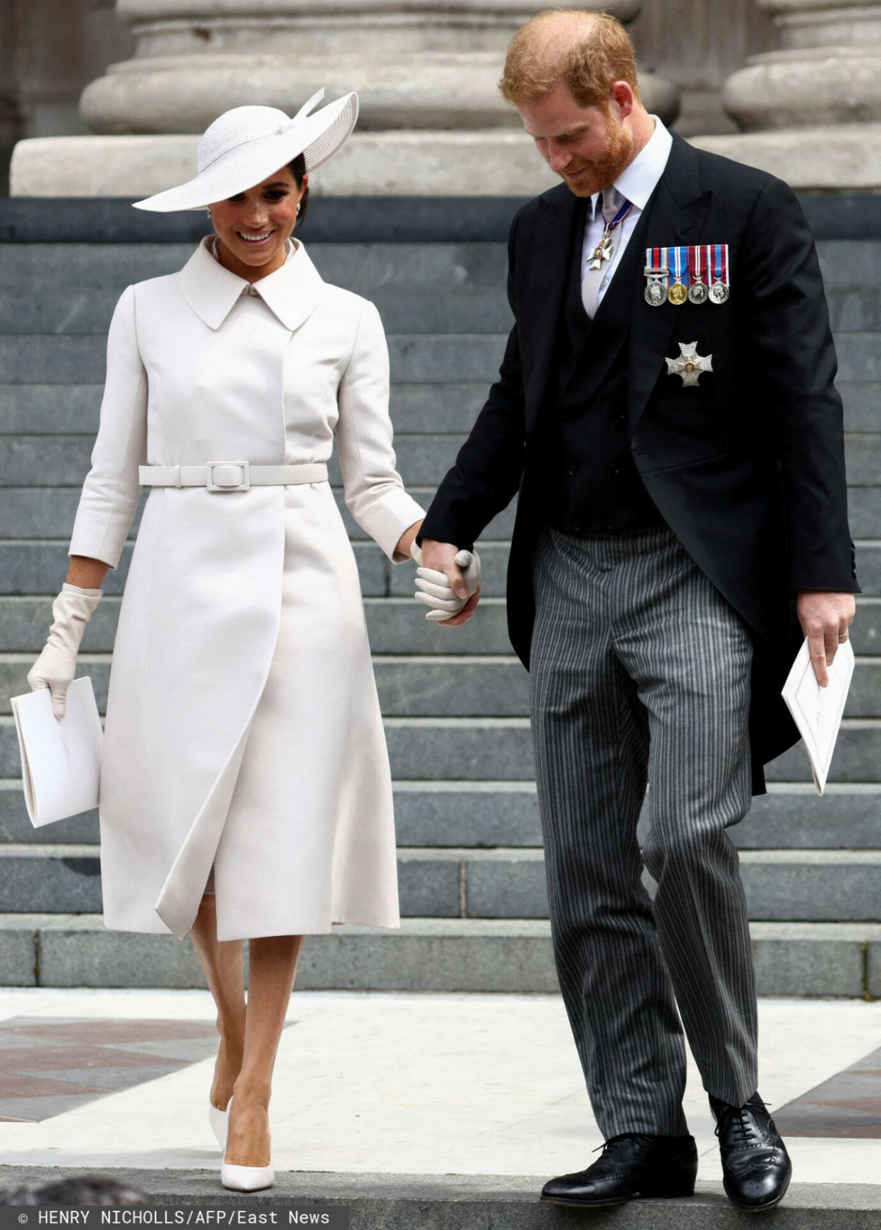 Meghan Markle w białej sukience idzie po schodach, trzymając za rękę księcia Harry'ego