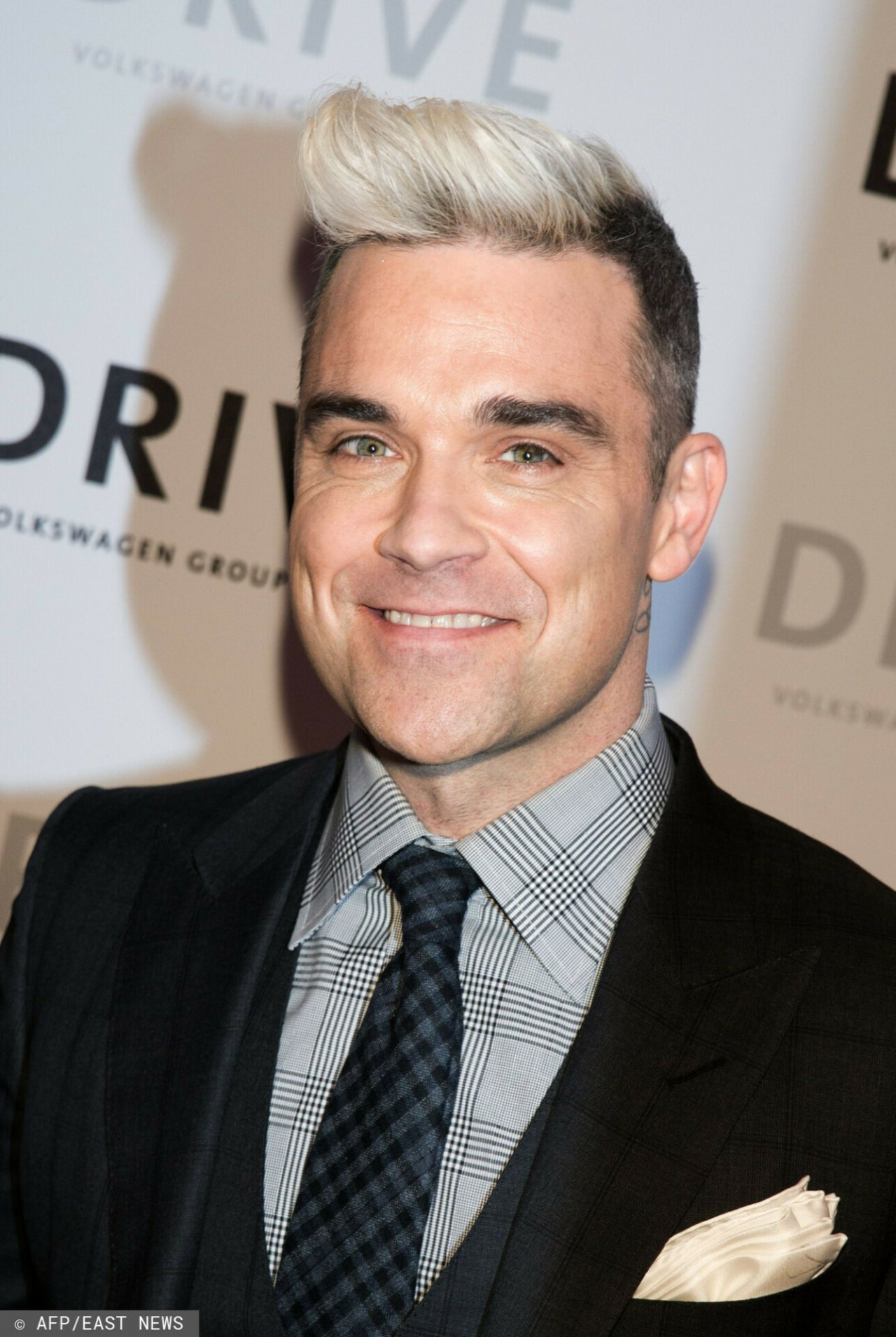 Robbie Williams z postawionymi włosami w kolorze blond i ciemnym garniturze