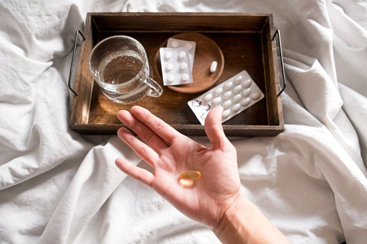Na jasnej pościeli drewniana taca z lekami i szklanką wody przysłonięta rozłożoną dłonią, na której są tabletki