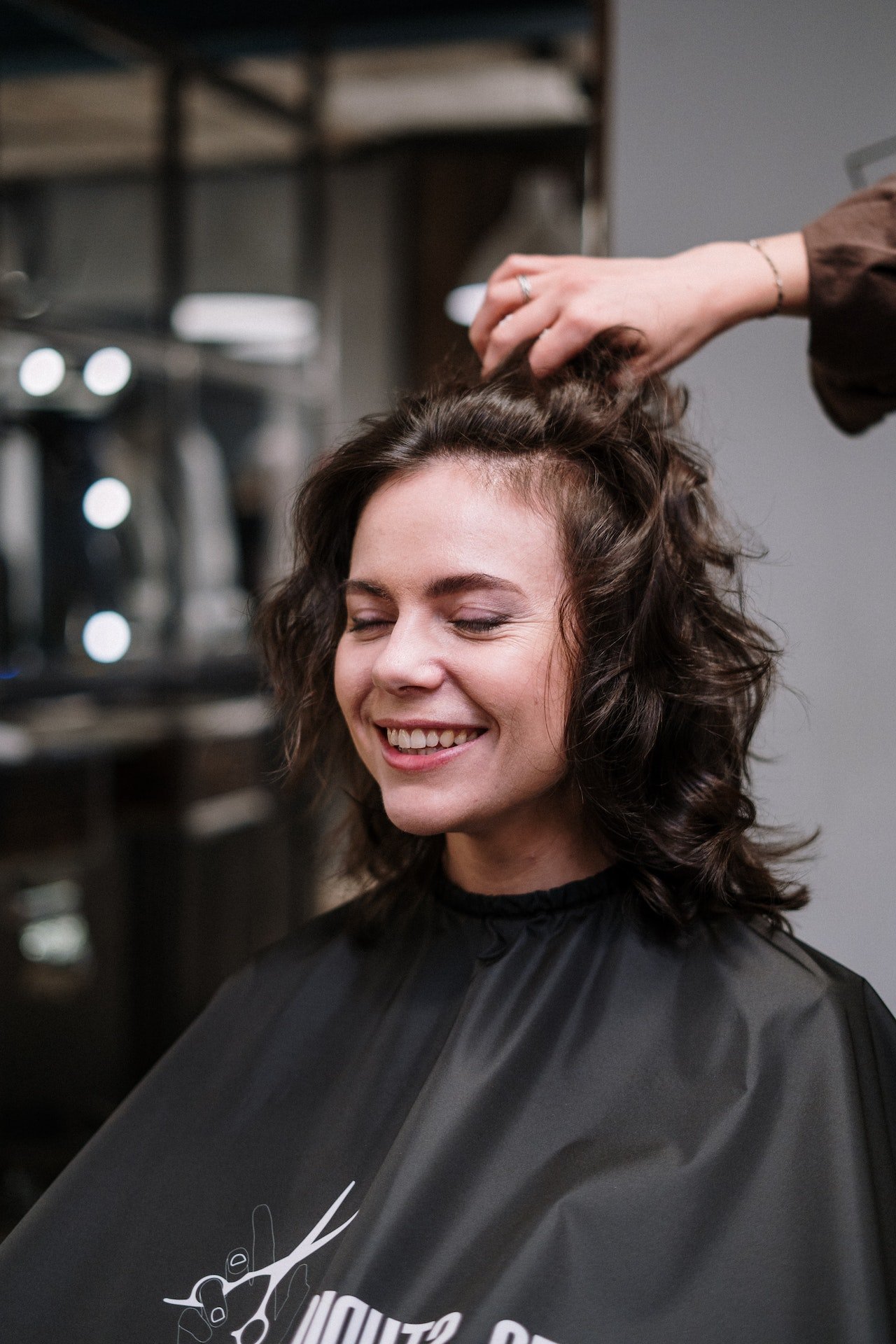 Radośnie uśmiechnięta kobieta w ciemnymi kręconymi włosach we fryzurze jak od fryzjera po użyciu Milk Shake z ręką fryzjera przeczesującą fryzurę
