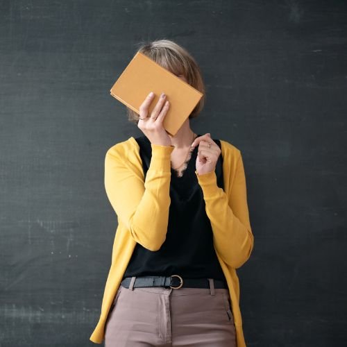 Nauczycielka w żółtym swetrze stoi pod tablicą, zasłania twarz notesem