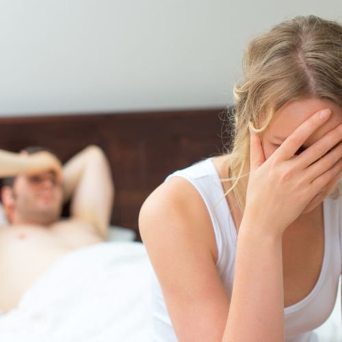 Kobieta siedzi na skraju łóżka z głową schowaną w dłoniach, w tle leżący w łóżku mężczyzna