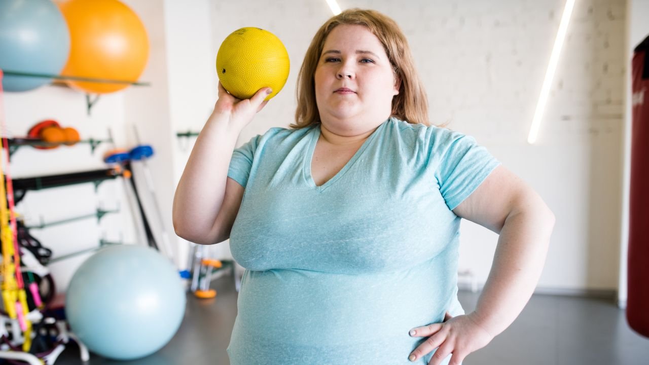 kobieta z nadwagą unosząca na siłowni żółtą piłkę 