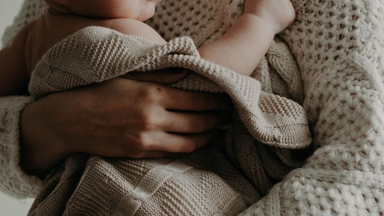 kobieta trzyma niemowlaka w ramionach, są okryci beżowym kocem