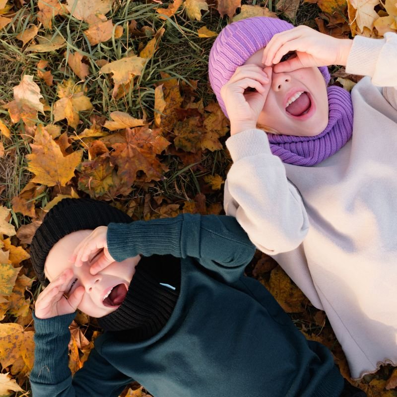 Dzieci w ubraniach z wełny merino gorpcore leżą na ziemi przykrej jesiennymi liśćmi robiąc śmieszne miny