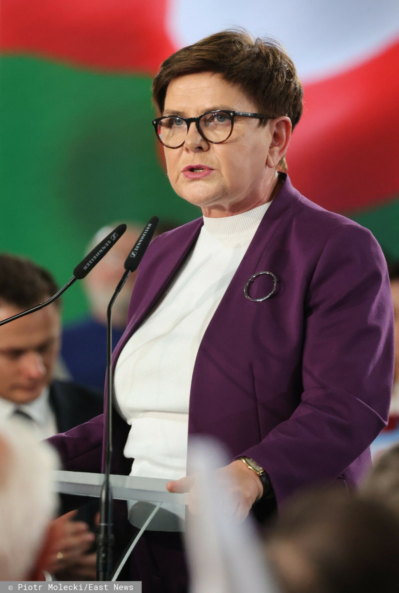 Beata Szydło w Białej bluzce, fioletowej marynarce z broszką przemawiająca do mikrofonu