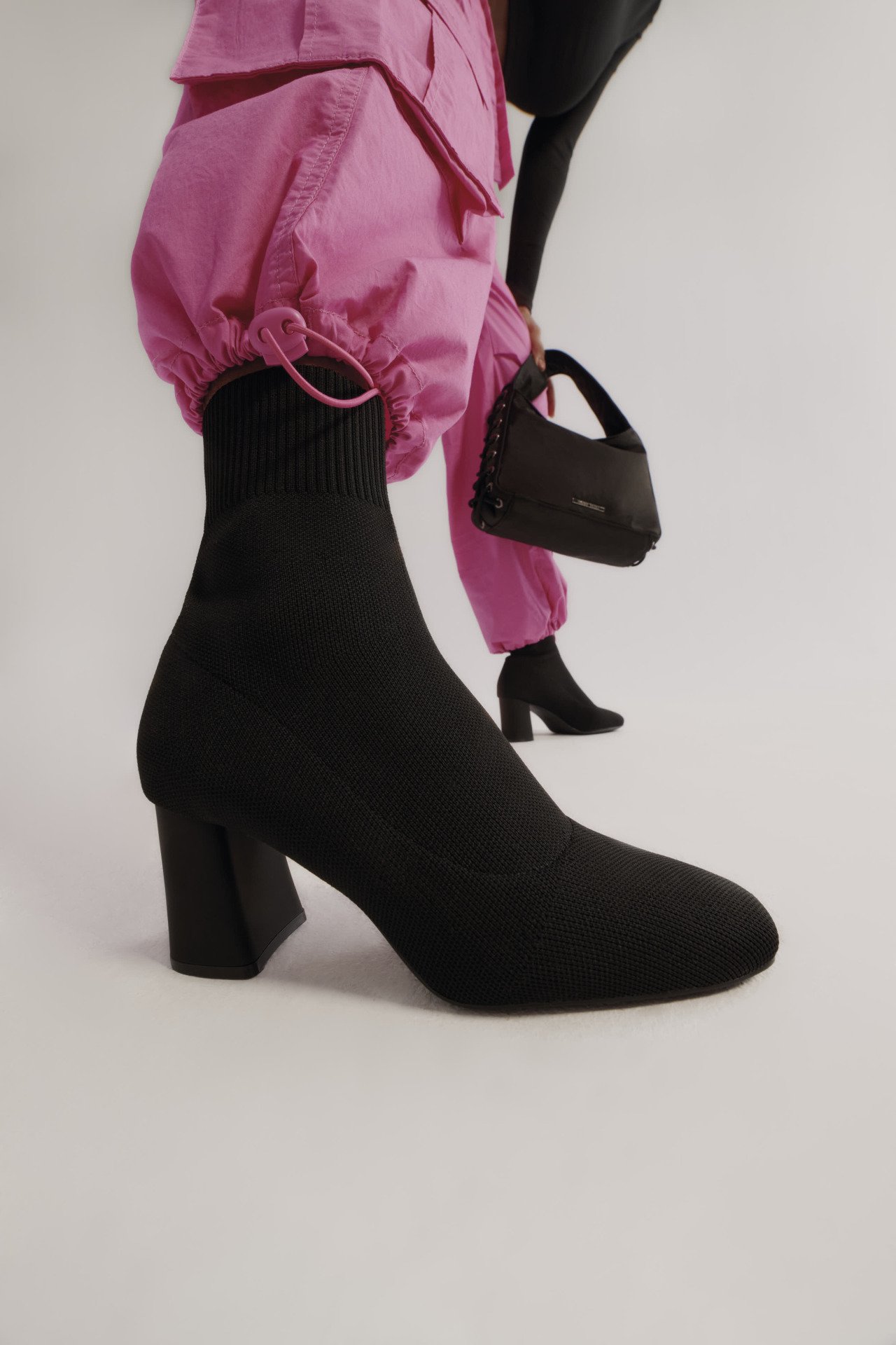 Kobieta w czarnych botkach na obcasie różowych spodniach z czarną torebką w ręku
