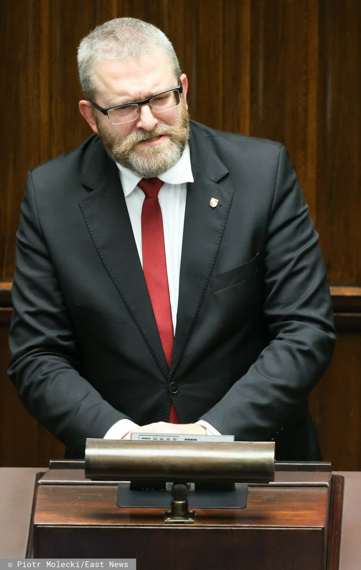 Grzegorz Braun w czarnym garniturze i z czerwonym krawatem
