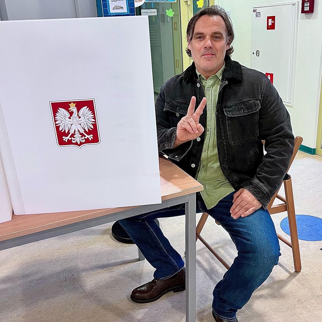 Jan Wieczorkowski w zielonej koszuli, skórzanej kurtce i jeansach w okręgu wyborczym podczas głosowania, pokazujący dłońmi znak pokoju