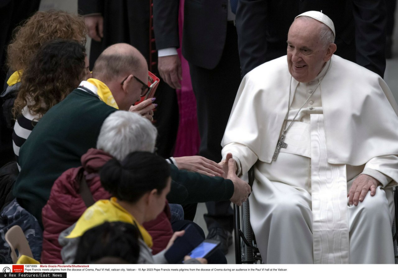 papież Franciszek w białej szacie uśmiechnięty wyciąga dłoń