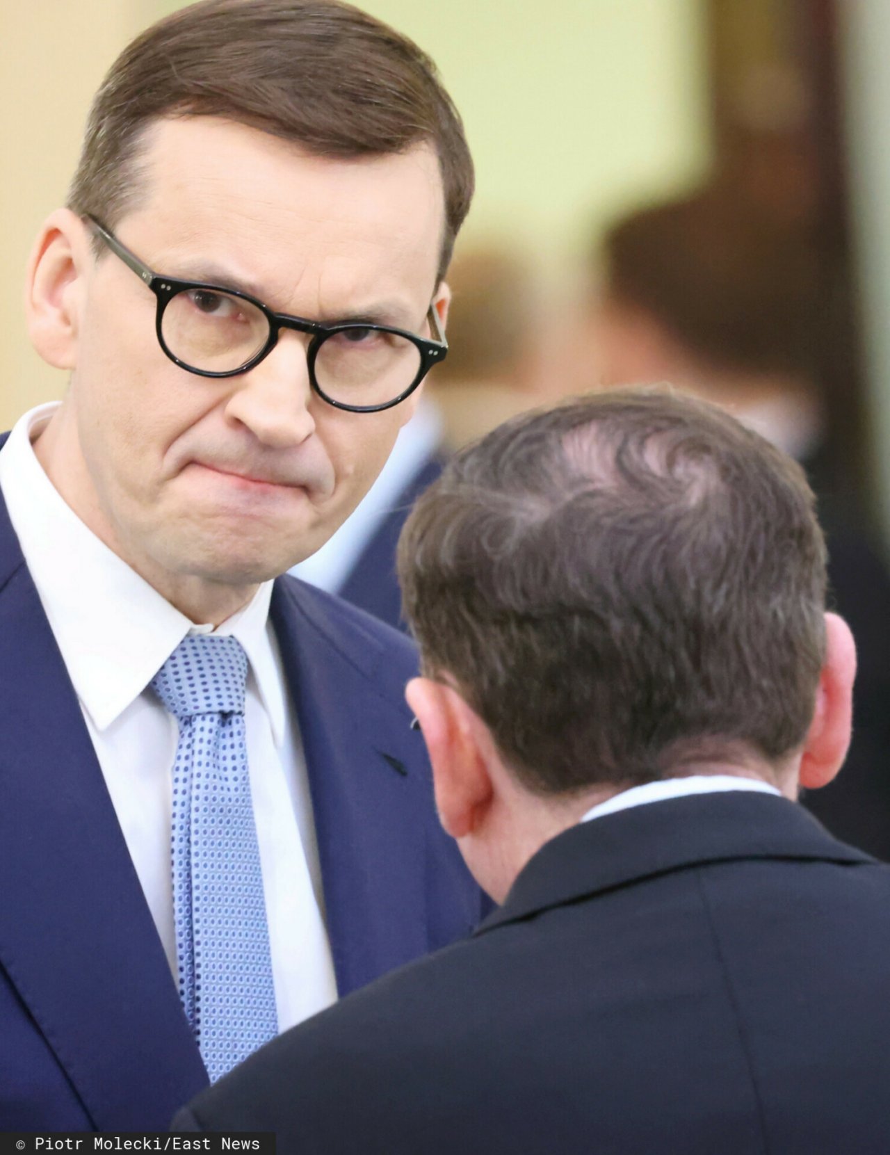 Mateusz Morawiecki w okularach i garniturze i Mariusz Kamiński stojący tyłem