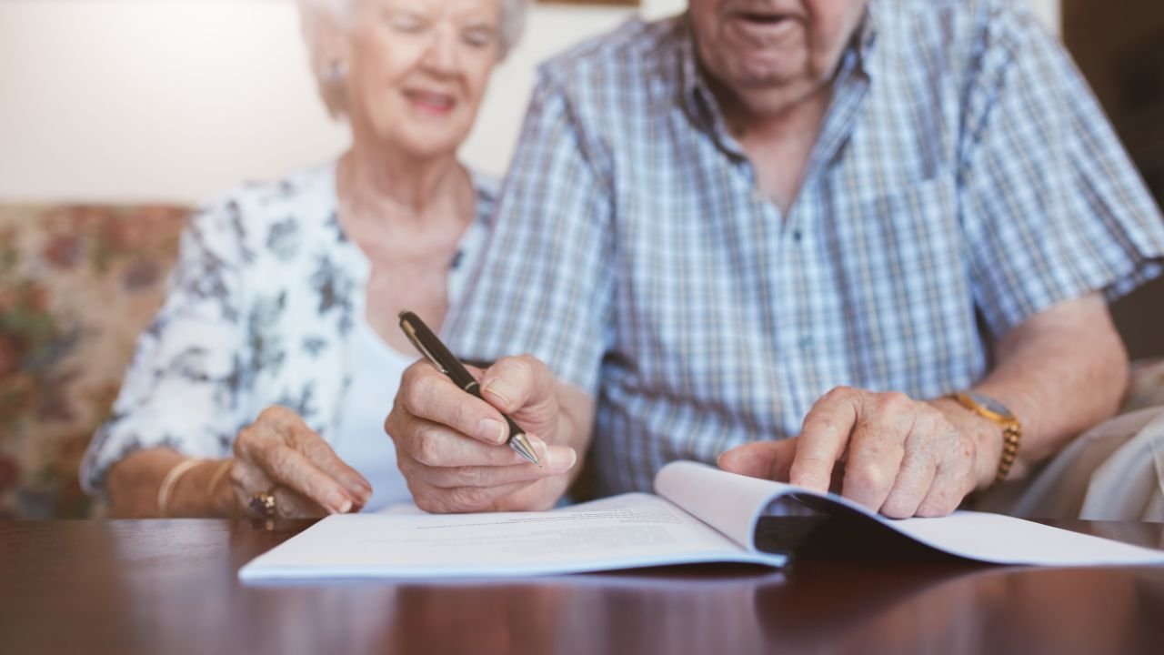 mężczyzna w podeszłym wieku ubrany w niebieską koszulę podpisujący dokument siedząc w obok kobiety ubranej w białą bluzkę z zielonymi wzorami 