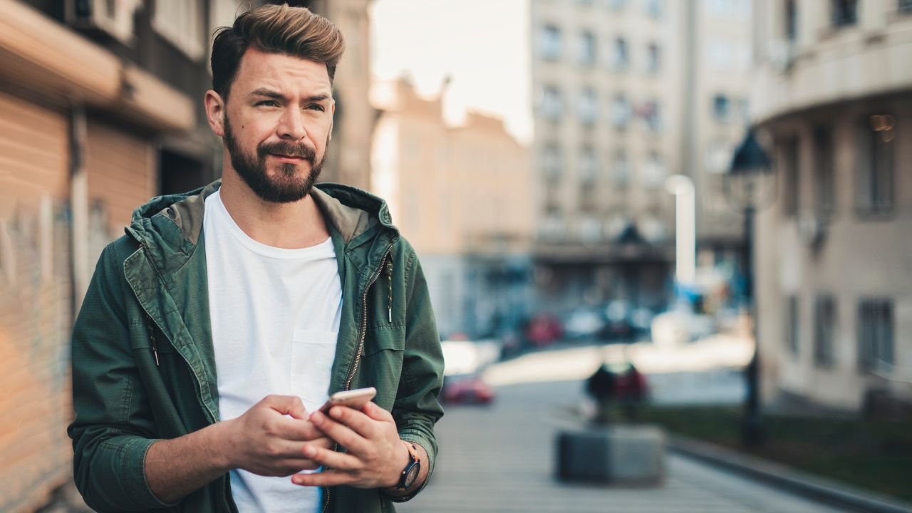 młody mężczyzna w białej koszulce i zielonej bluzie trzymający w ręku telefon komórkowy 