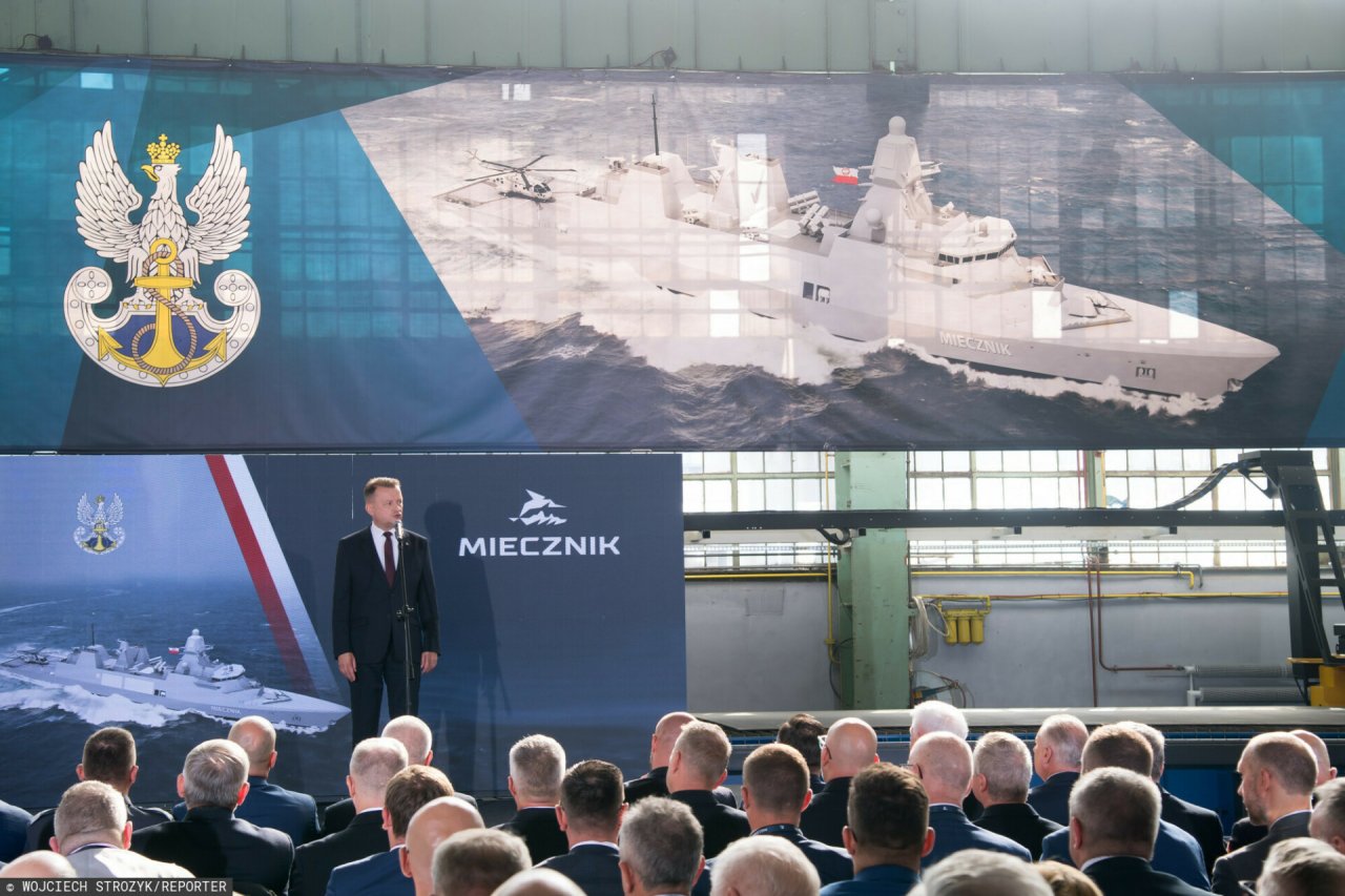 Mariusz Błaszczak stoi na tle plakatu z okrętem Miecznik