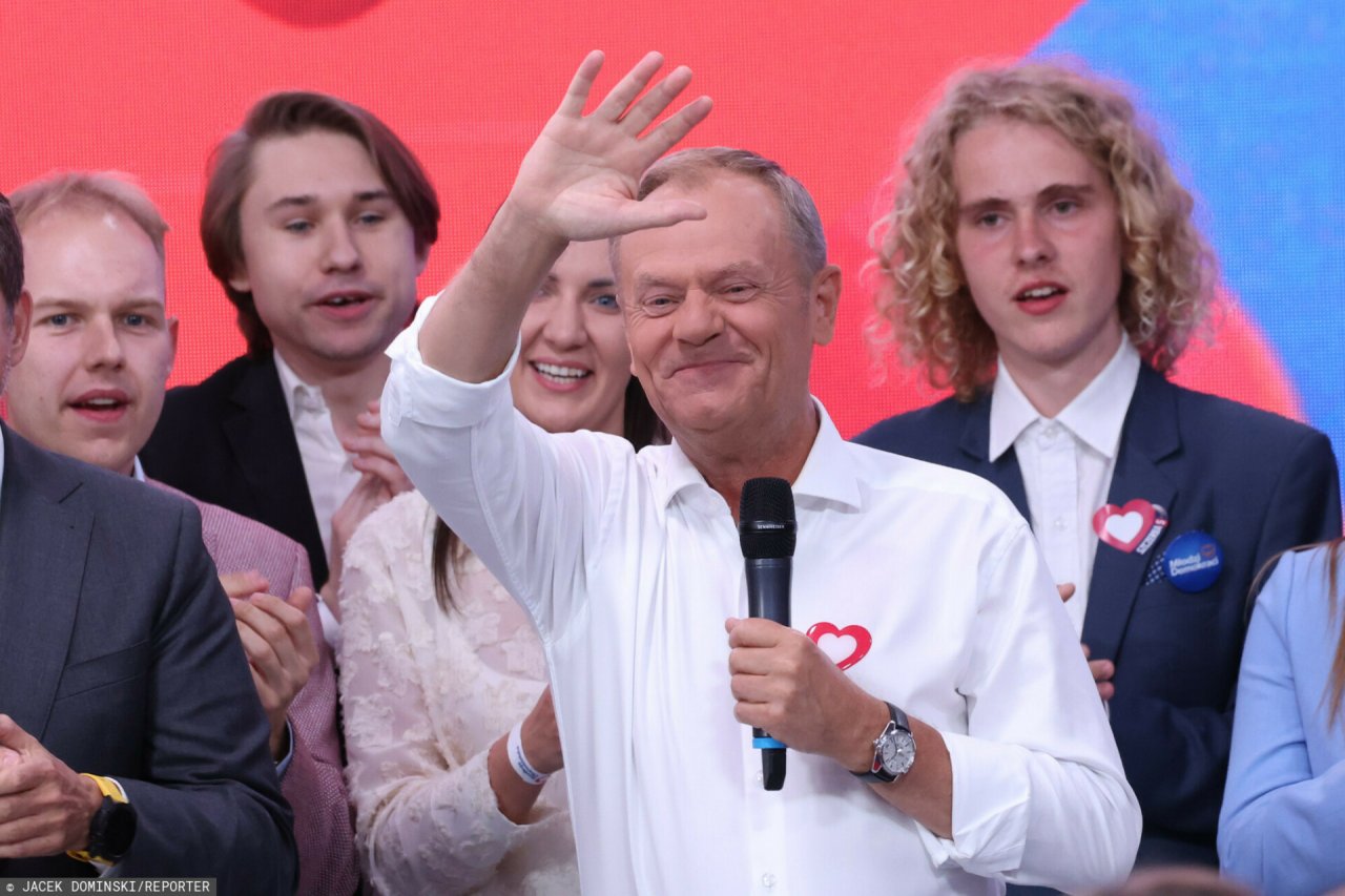 Uśmiechnięty Donald Tusk w białej koszuli podnosi rękę do góry
