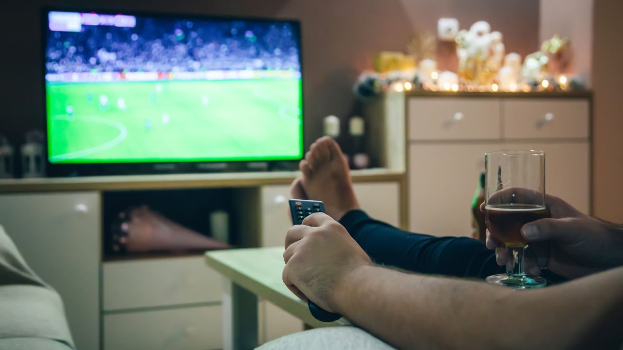 mężczyzna w niebieskich spodniach siedzący na kanapie z pilotem w dłoni przed telewizorem na którym wyświetlany jest mecz piłki nożnej 