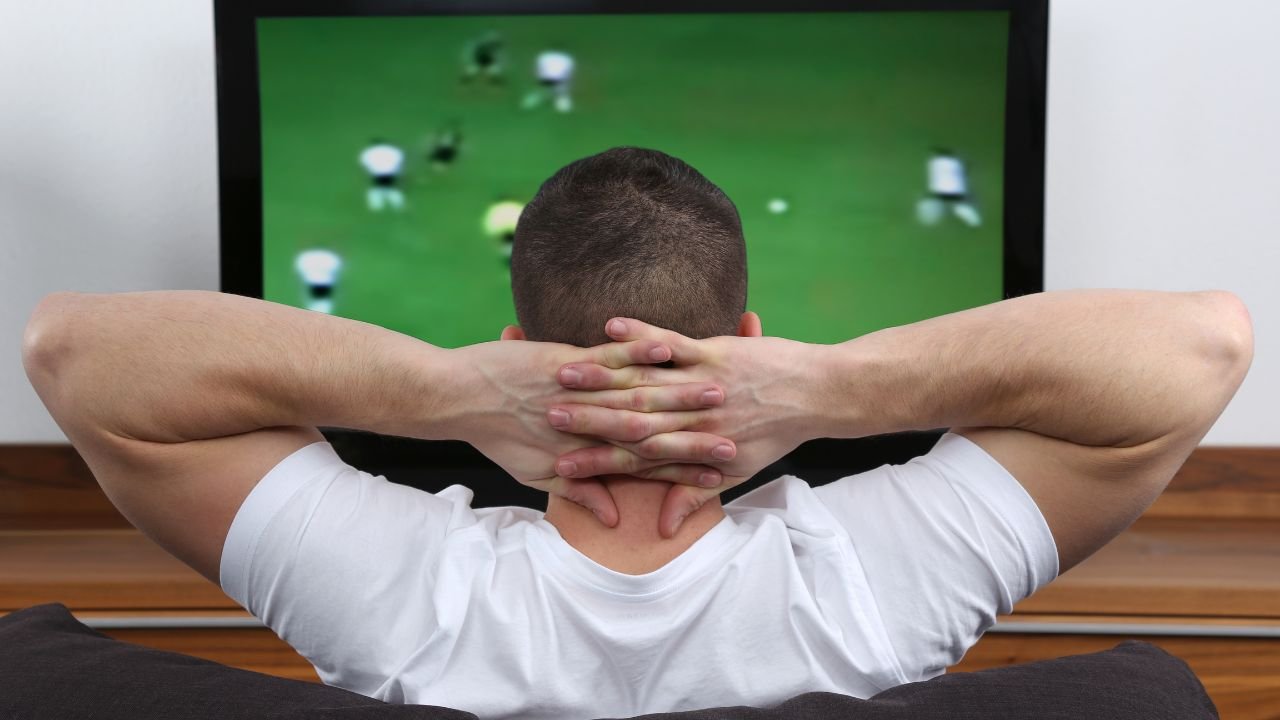 mężczyzna w białej koszulce z założonymi rękoma za szyję siedzący przed telewizorem na którym wyświetlany jest mecz piłki nożnej 