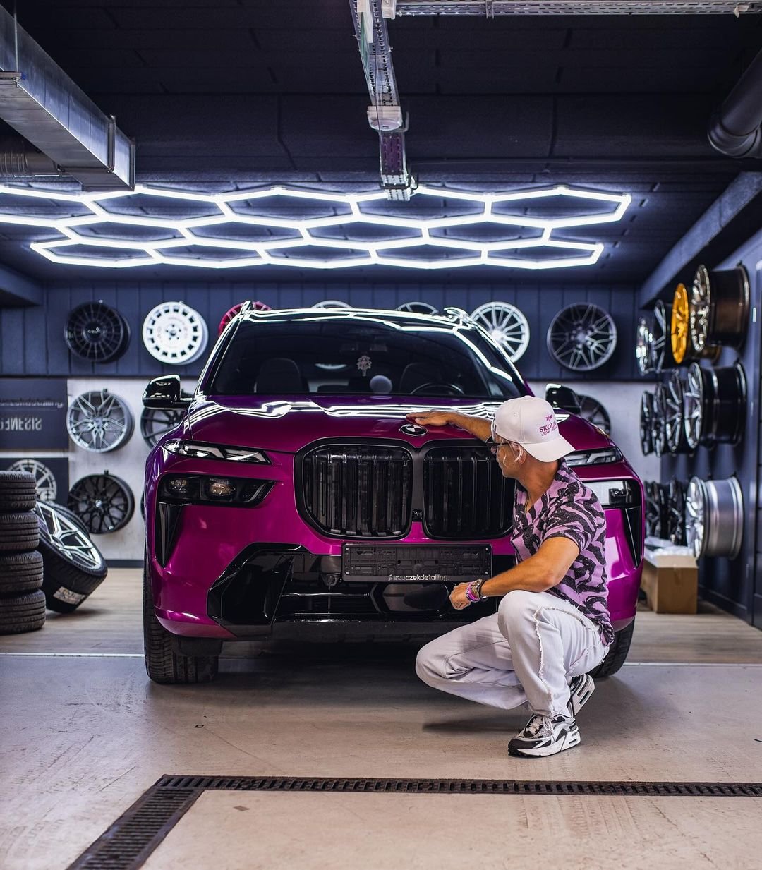 Skolim w fioletowej koszuli w pasy i jasnych spodniach kuca przed różowym BMW w garażu