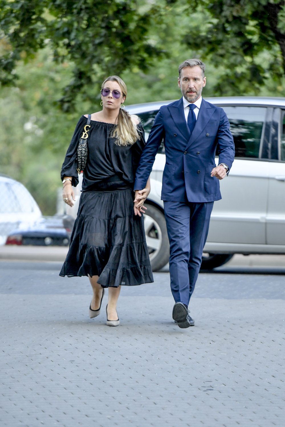 Karolina Ferenstein-Kraśko w czarnej długiej sukni i Piotr Kraśko w granatowym garniturze idą na tle auta i zieleni