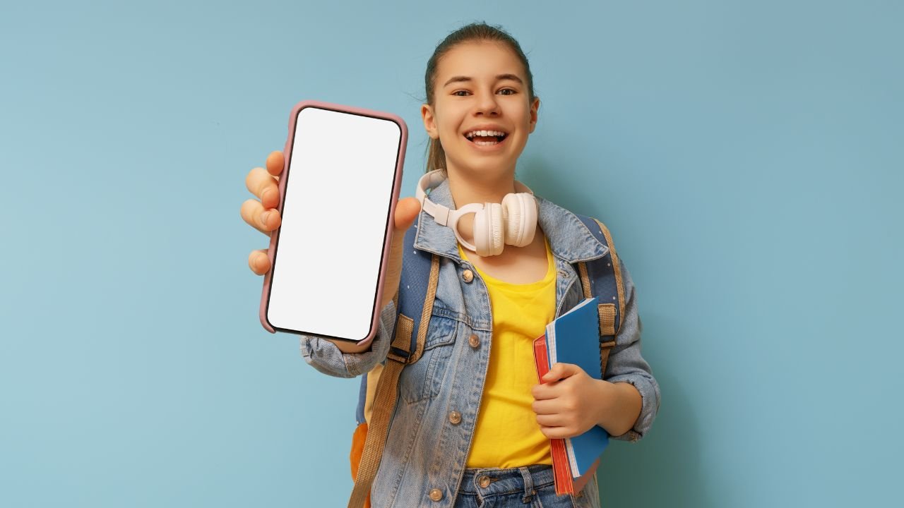 Uśmiechnięta dziewczynka ze słuchawkami na szyi wyciąga w ręce telefon