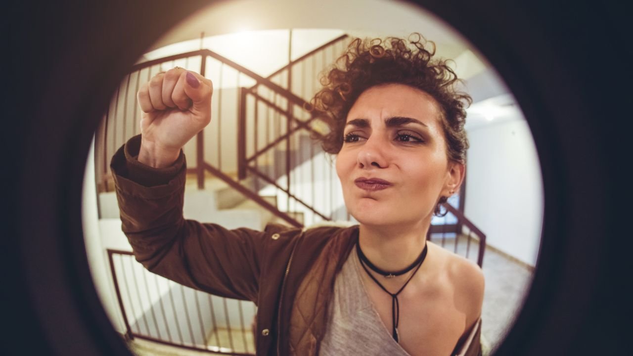 Widziana przez wizjer kobieta puka w drzwi z niezadowoloną miną