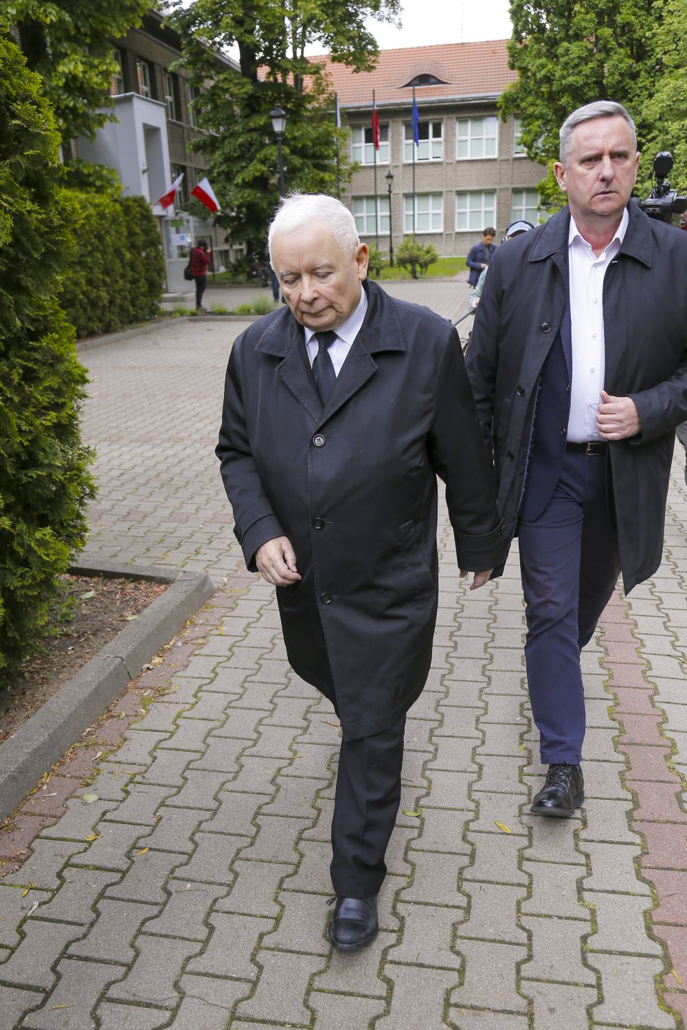 Jarosław Kaczyński podąża na wybory prezydenckie w czarnym płaszczu, białej koszuli i czarnym krawacie w towarzystwie wysokiego, eleganckiego mężczyzny 
