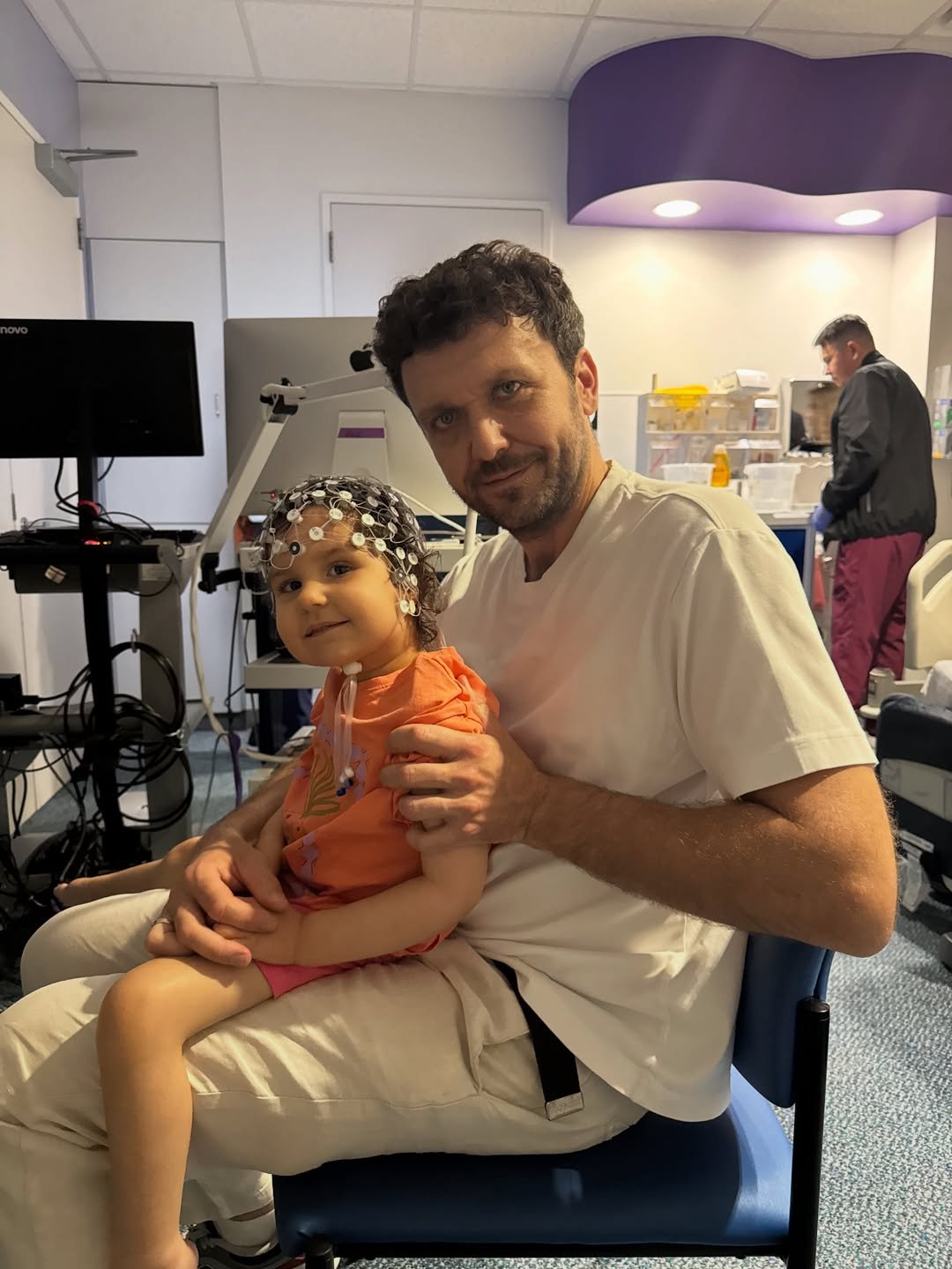 Bartek Marszałek w białym T-shircie trzyma na kolanach córkę podczas badania EEG