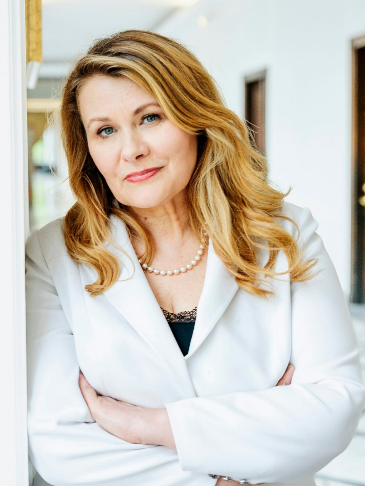 Katarzyna Piekarska w czarnym topie i białej marynarce opiera się o ścianę