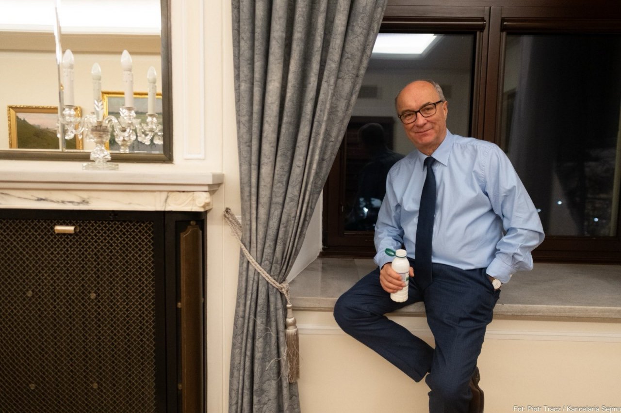 Włodzimierz Czarzasty w niebieskiej koszuli siedzi na parapecie okna pijąc kefir