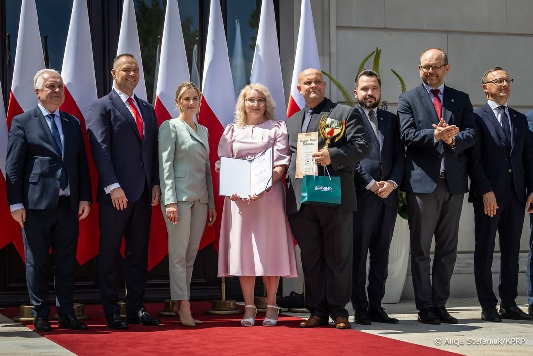 Marta Nawrocka w oliwkowym komplecie pozuje z kilkoma osabami na tle flag Polski