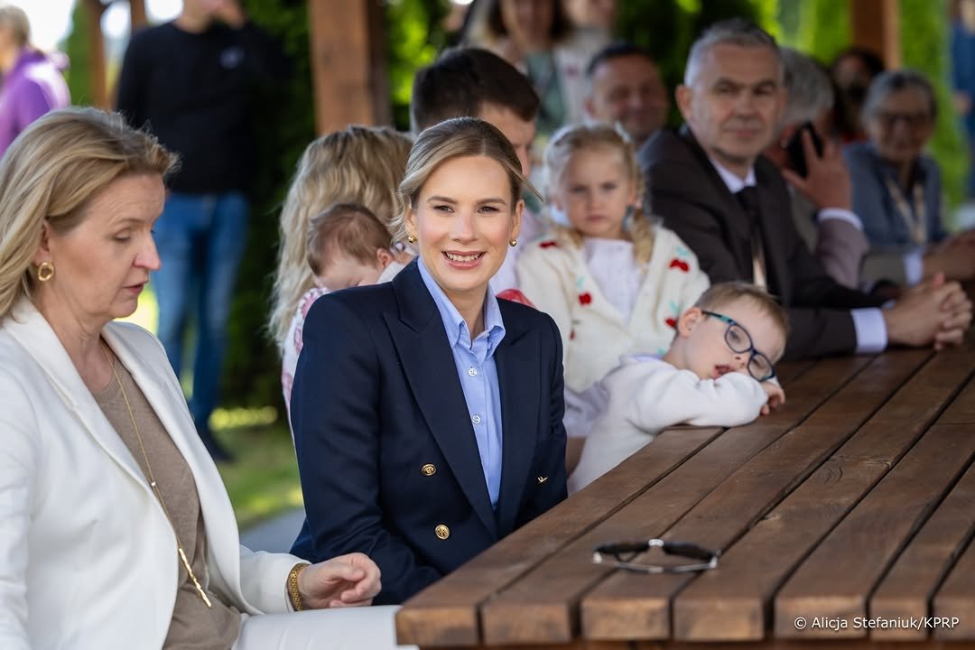 uśmiechnięta Marta Nawrocka siedzi przy stole, w tle grubka osób
