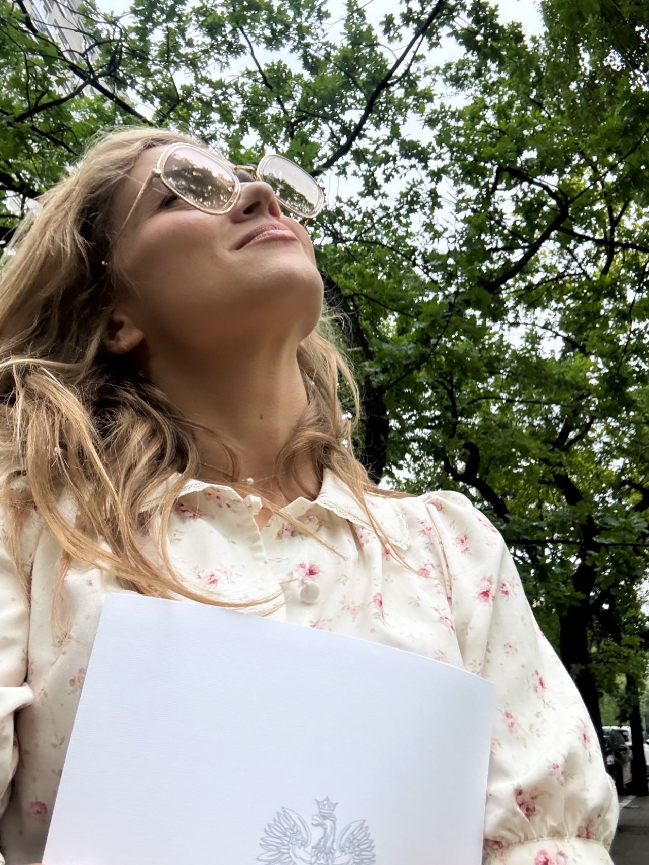 ubrana w jasną koszule agnieszka kaczorowska spogląda w górę, stojąc wśród drzew