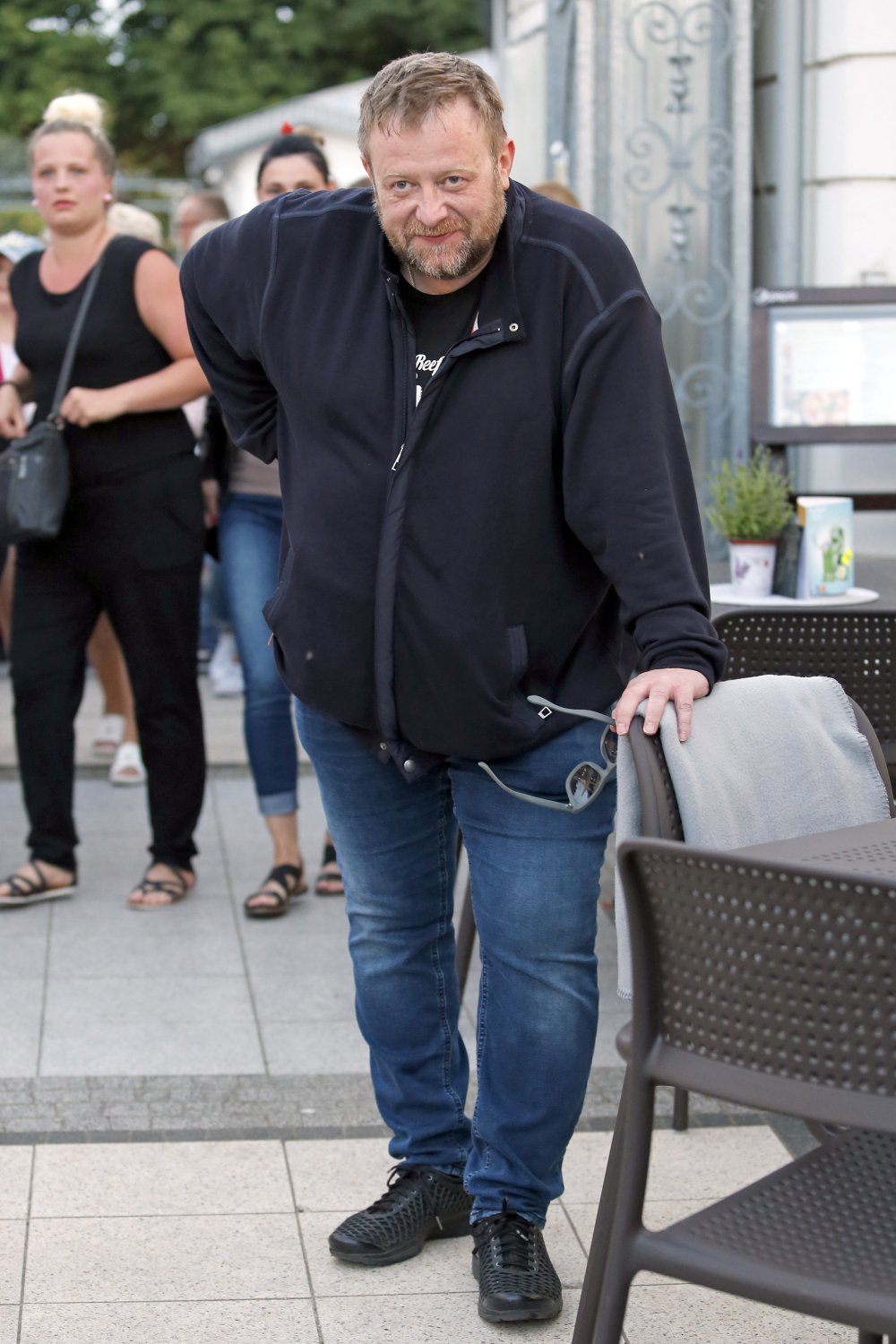 Olaf Lubaszenko w ciemnej kurtce i jeansach posbiera sie o fotel stojąc obok grupki osób